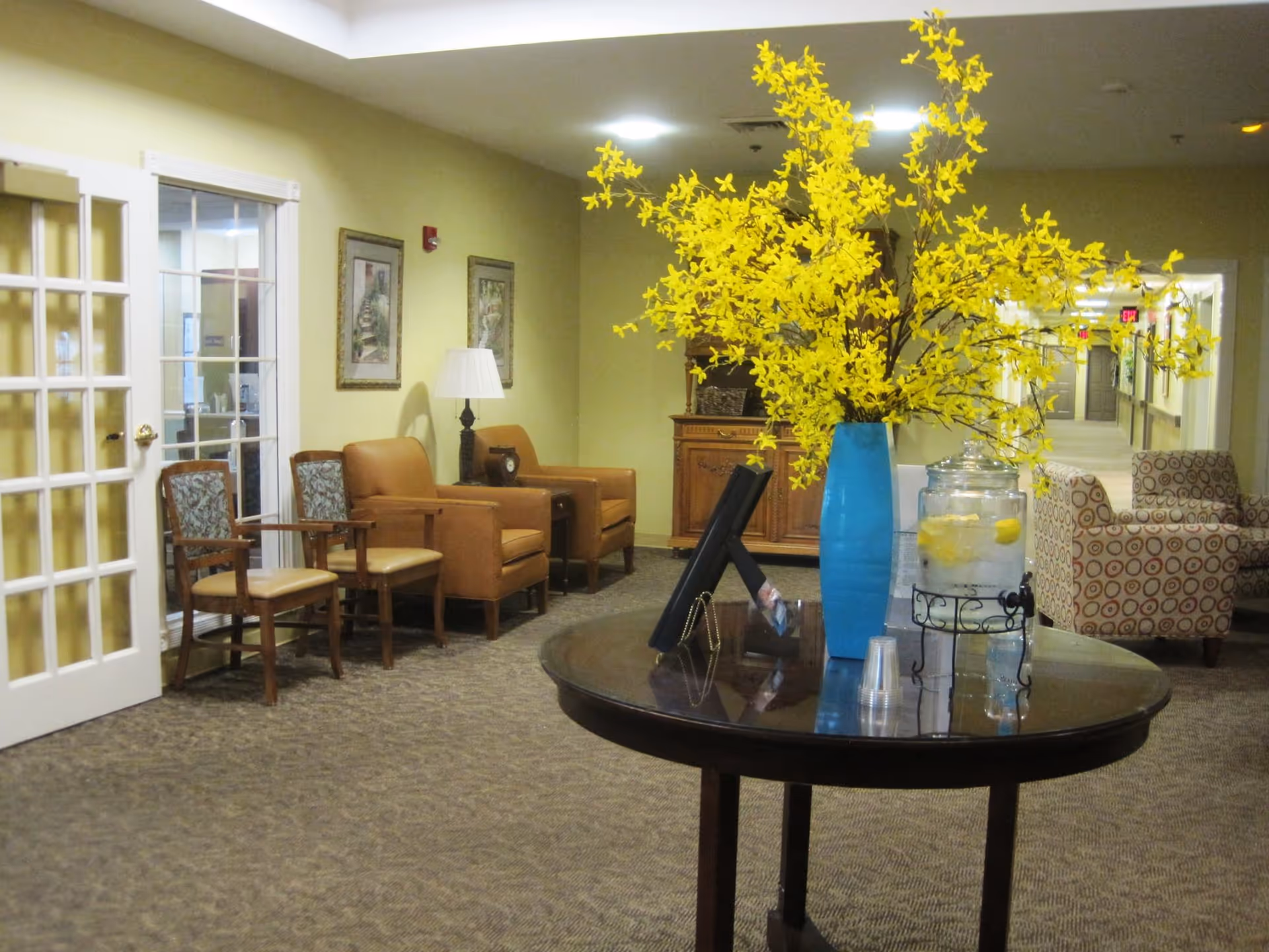 A senior living facility common area with a round table in the foreground holding a blue vase with bright yellow flowers and a glass dispenser filled with water and lemon slices. The background shows a hallway with patterned armchairs, leather chairs, framed artwork on the walls, a lamp, and a wooden cabinet.