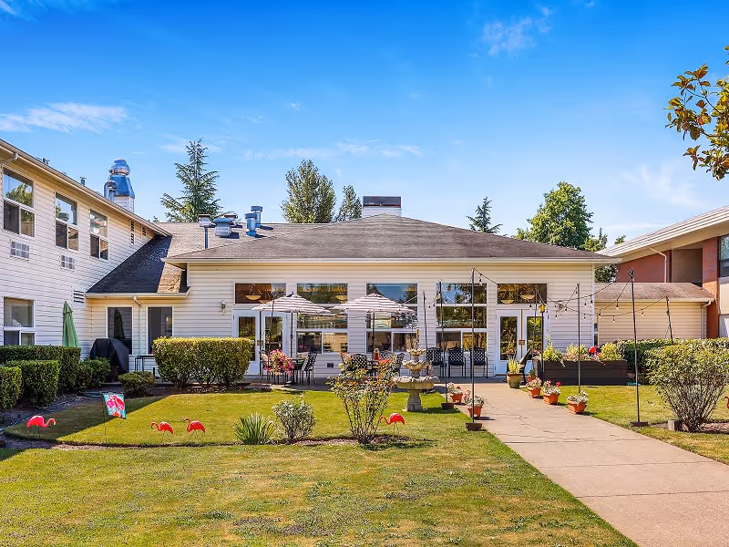 Exterior view of White River Assisted Living facility showing a single-story building with large windows, outdoor seating with umbrellas, a fountain, and a well-maintained lawn with decorative flamingos and potted plants under a clear blue sky.
