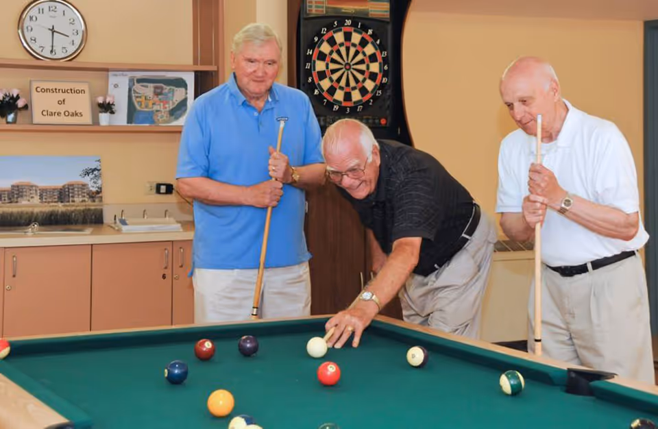 Three elderly men playing pool in a recreational room. One man is taking a shot while the other two watch, each holding a pool cue. Behind them is a dartboard, a clock on the wall, and some pictures and signs on a shelf.