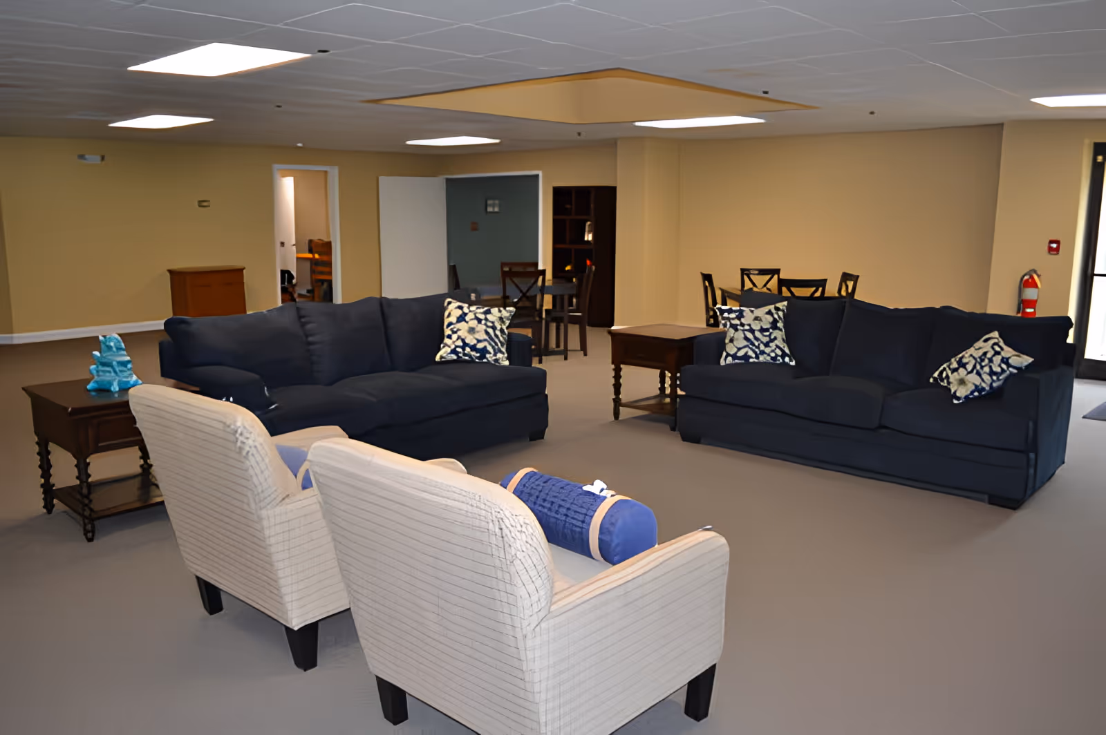 A spacious senior living common area with two dark blue sofas adorned with patterned pillows, two light-colored armchairs with a blue bolster pillow, wooden side tables, and a dining table with chairs in the background. The room has beige walls, a gray carpet, and recessed ceiling lights.