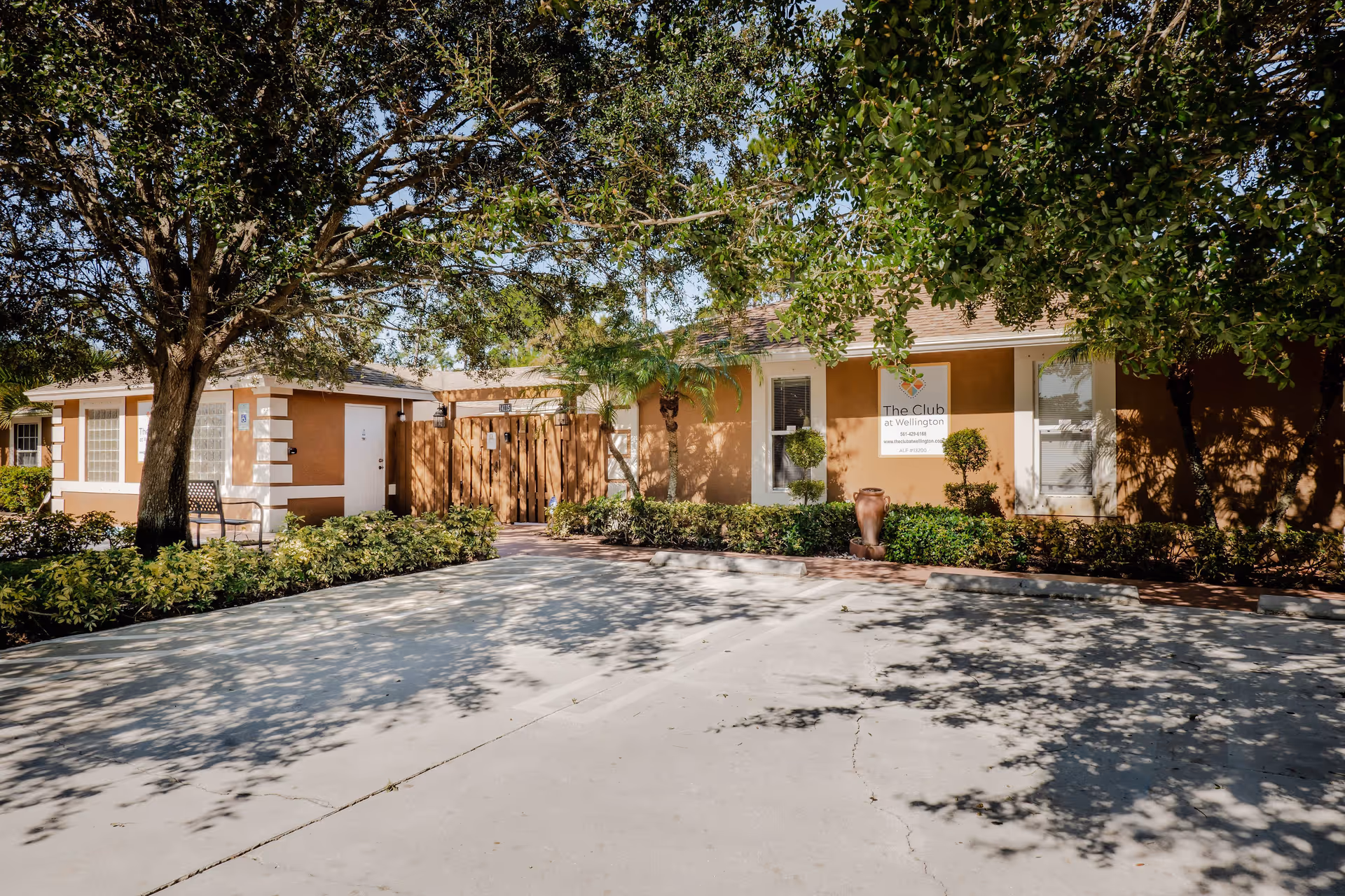 Exterior view of The Club at Wellington assisted living and memory care facility showing a single-story building with tan walls and white trim. There are trees and shrubs in front of the building, a wooden gate, and a parking area with shadows of tree branches on the pavement.