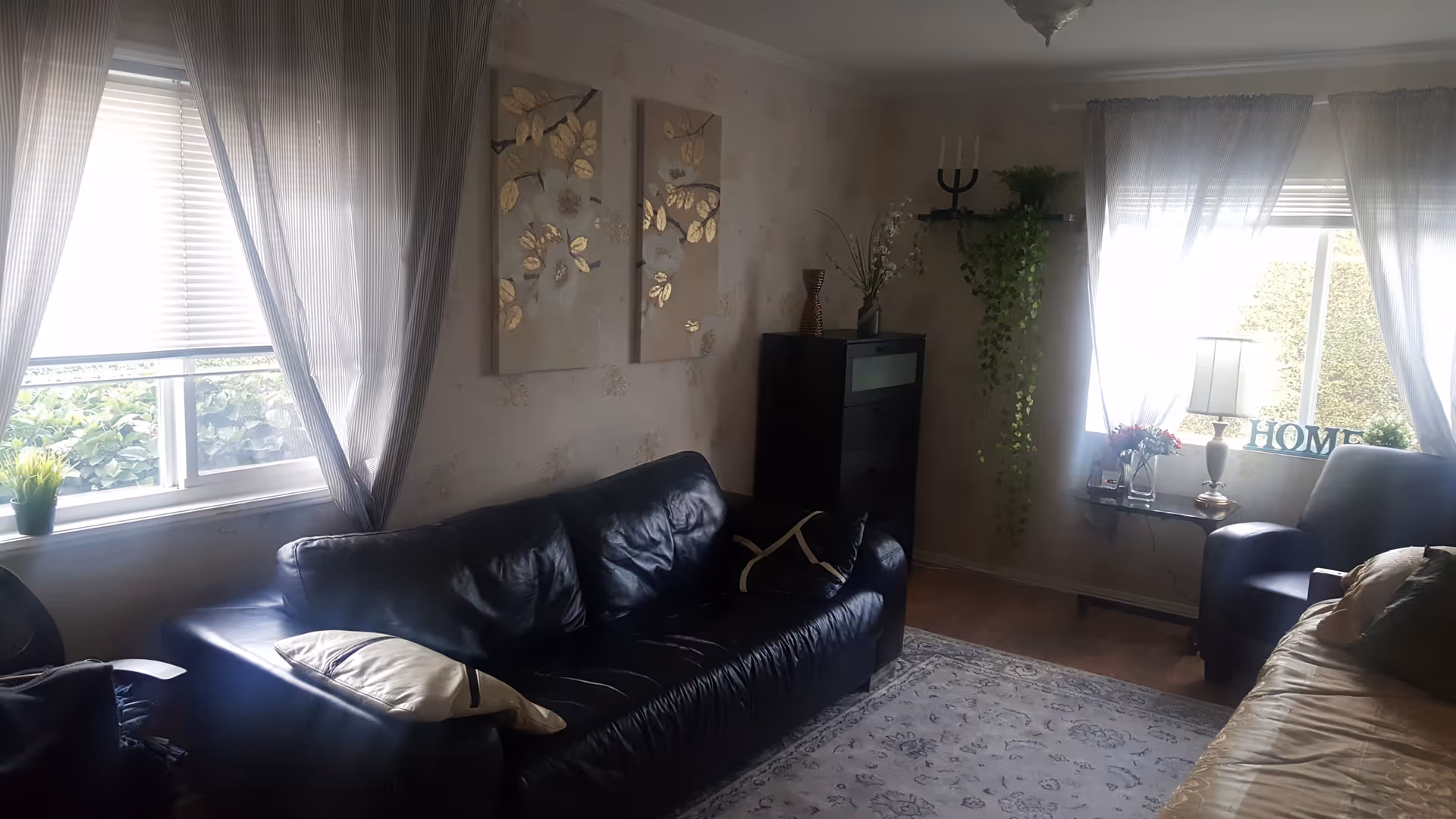 A cozy living room with a black leather sofa and a beige sofa, both with cushions. Two windows with sheer curtains allow natural light to brighten the room. There is a small table with a lamp, flowers, and a decorative 'HOME' sign. The walls have floral artwork and a tall black cabinet with decorative items on top. A patterned rug covers the wooden floor.