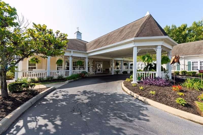 Front exterior view of a senior living facility with a covered driveway entrance, white railings, hanging plants, and well-maintained landscaping including trees and flower beds under a clear blue sky.