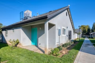 Exterior view of a single-story residential building with a light gray stucco finish and a teal front door. The building has a sloped roof with an HVAC unit on top. There is a concrete walkway alongside the building, bordered by grass and small plants. Other similar buildings and trees are visible in the background under a clear blue sky.