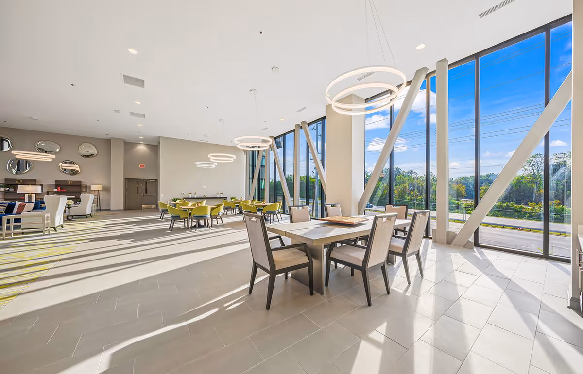 Bright and spacious interior room with large floor-to-ceiling windows allowing natural light to flood in. The room features modern circular hanging light fixtures, a dining table with six chairs in the foreground, and several smaller tables with green chairs in the background. The walls are light-colored, and there are decorative mirrors and lamps on the left side of the room.