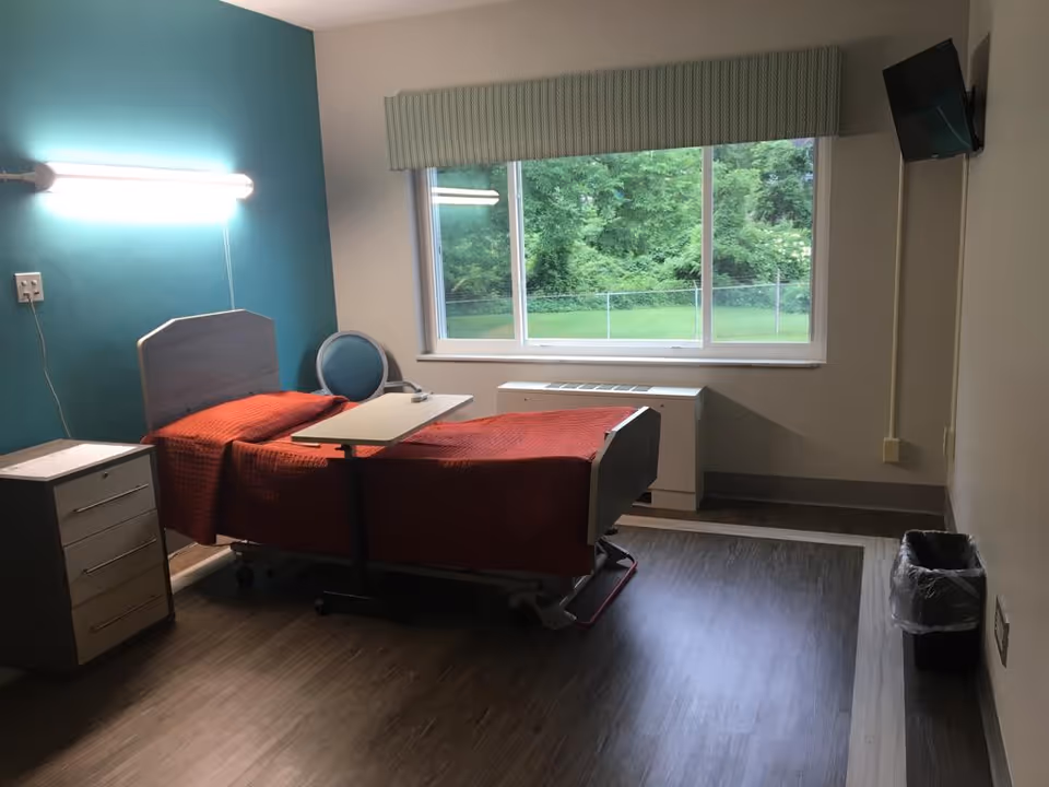 A simple nursing facility room with a single hospital bed covered in a red blanket, a small overbed table, a blue chair, a bedside cabinet with drawers, a wall-mounted fluorescent light, a large window showing green trees outside, a wall-mounted TV, and a trash bin on the floor.