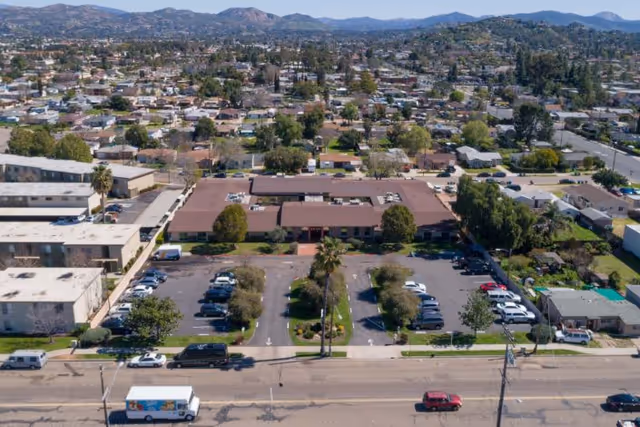 Aerial view of a single-story building with a large parking lot in front, surrounded by residential houses and trees, with mountains visible in the background under a clear sky.