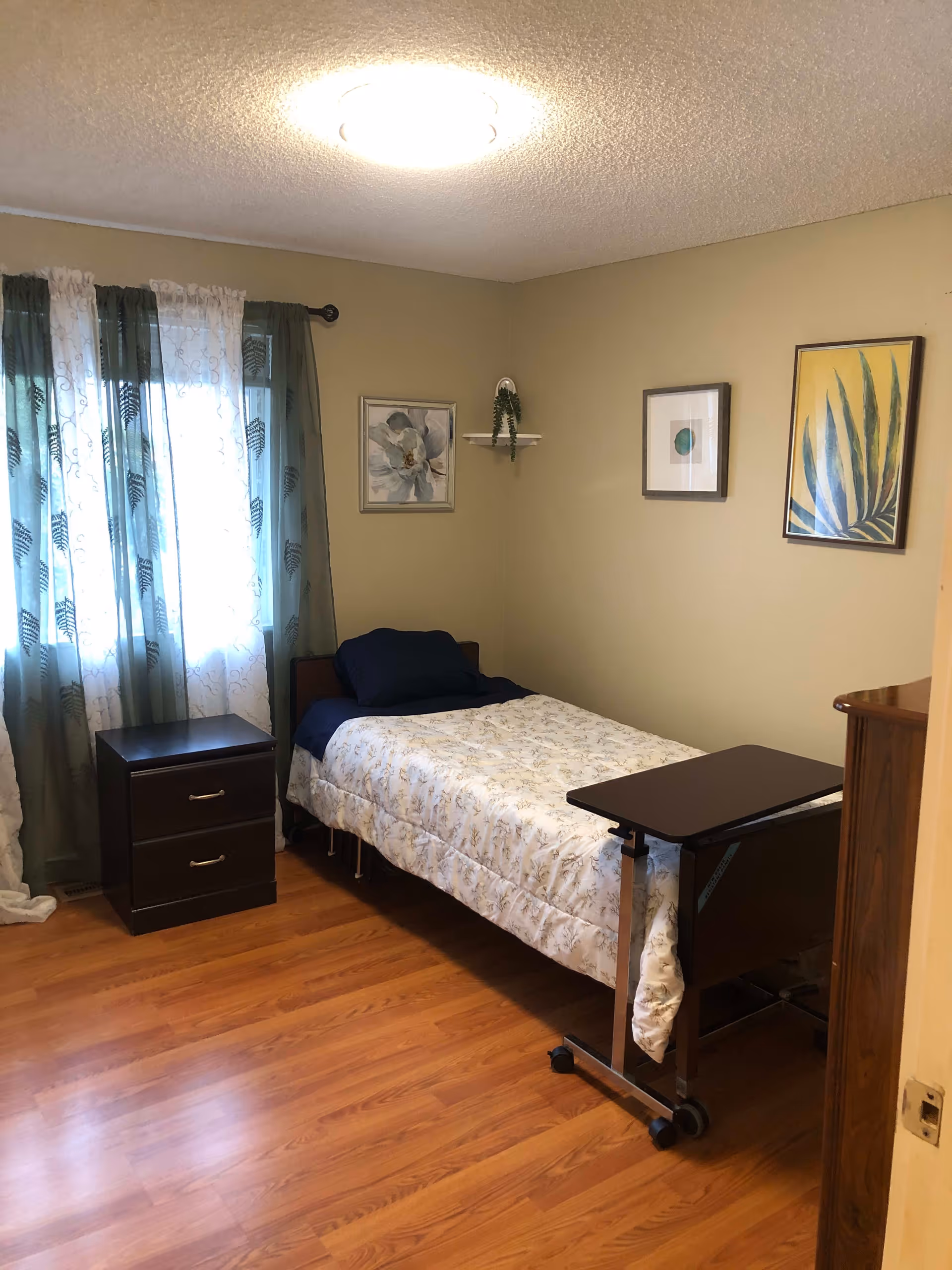 A small bedroom with a single bed covered in a floral comforter and dark blue pillow. Next to the bed is a dark wooden nightstand with two drawers. A rolling overbed table is positioned at the foot of the bed. The room has light brown walls with three framed pictures hanging on them and a small white shelf with a green plant. A window with sheer white and green curtains lets in natural light. The floor is wood laminate and a round ceiling light illuminates the room.