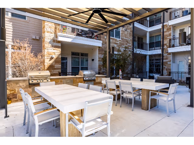 Outdoor patio area with two large rectangular tables surrounded by white chairs under a pergola. There are two stainless steel grills built into a stone counter against the building wall. The building has stone and siding exterior with balconies and windows overlooking the patio.