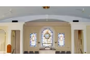 Interior view of a chapel or worship area with three stained glass windows above an altar. The room has white walls, columns, and ceiling fans, with chairs arranged in rows facing the altar.