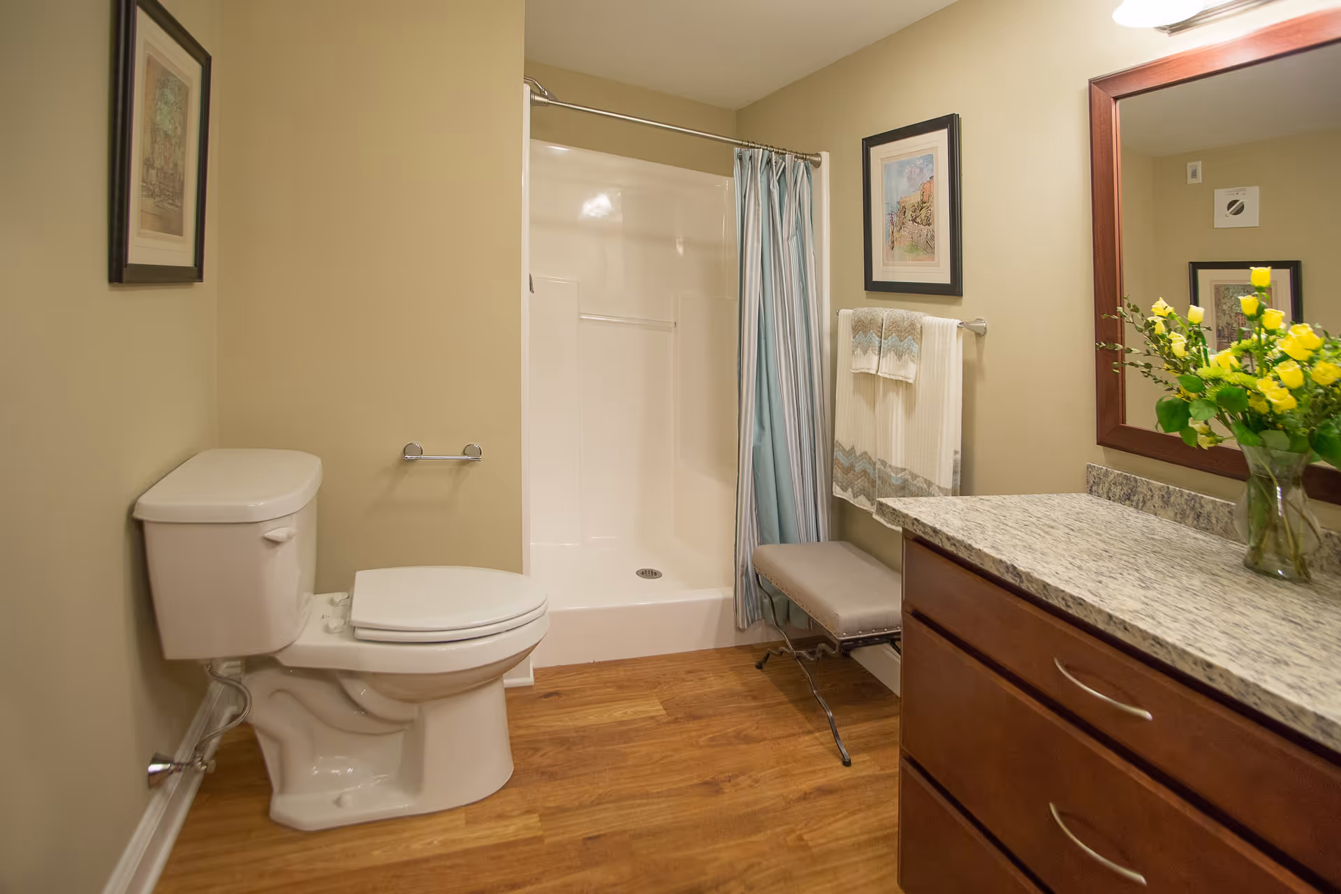 A clean bathroom with a white toilet, a walk-in shower with a blue shower curtain, a wooden vanity with a granite countertop, a large mirror, a vase with yellow flowers, and framed artwork on the walls. There is also a small cushioned bench and towels hanging on a rack.