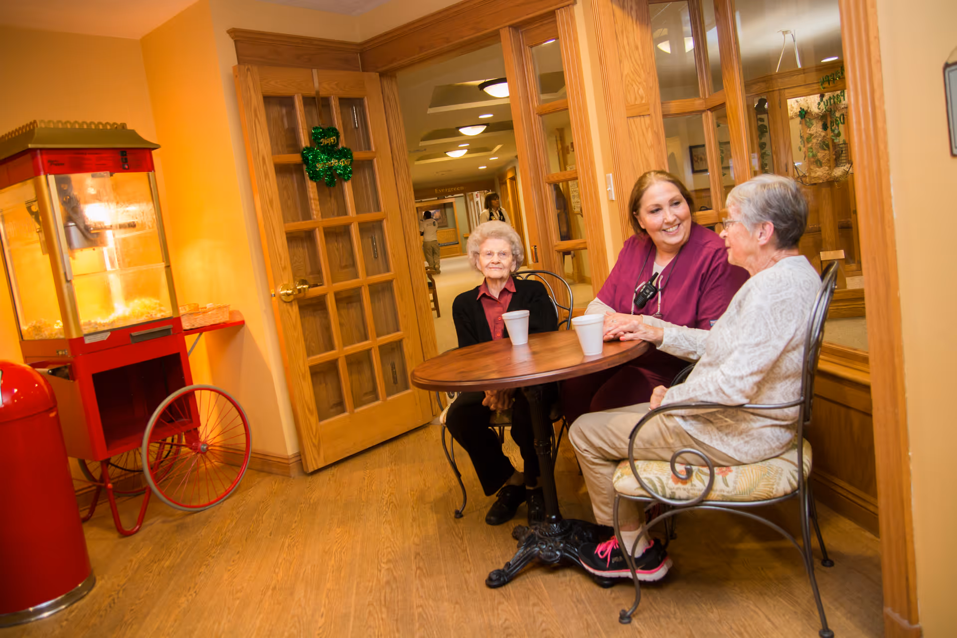 Two elderly women and a caregiver sitting around a small round table in a cozy indoor setting. The caregiver is smiling and engaging with one of the elderly women. There is a popcorn machine on the left side and wooden doors with glass panels in the background.