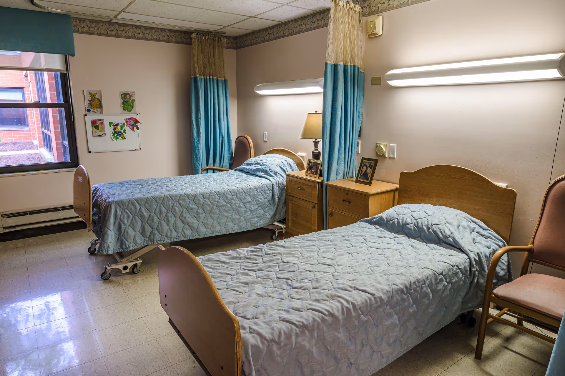 A shared room in a healthcare facility with two single beds covered in light blue quilts. Each bed has a wooden headboard and footboard. Between the beds are two wooden nightstands with framed photos and a lamp. Blue and beige privacy curtains hang on the wall behind the beds. There is a window on the left side with a green valance, and a chair is positioned near the bed on the right. The floor is tiled, and the walls are painted light beige.