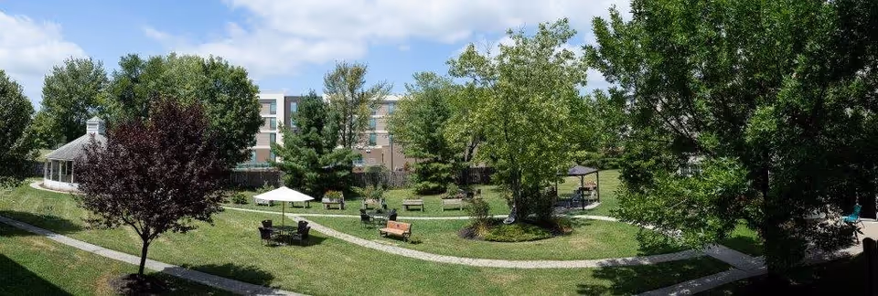 A spacious outdoor garden area with green grass, several trees, benches, and tables with umbrellas. There is a gazebo on the left side and a multi-story building in the background under a partly cloudy blue sky.