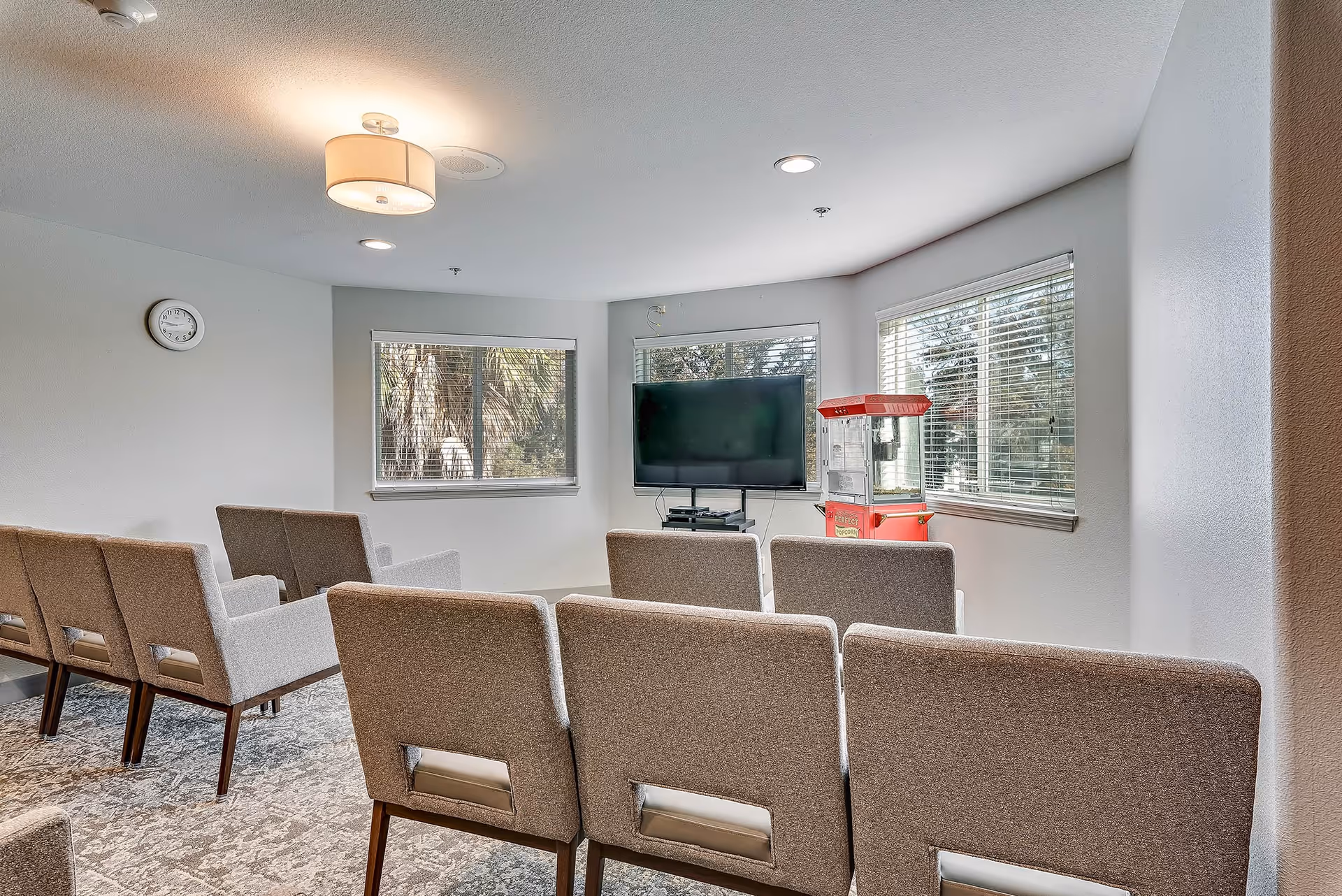 Rows of upholstered chairs arranged theater-style facing a wall-mounted TV and a popcorn machine in a bright common room with windows.