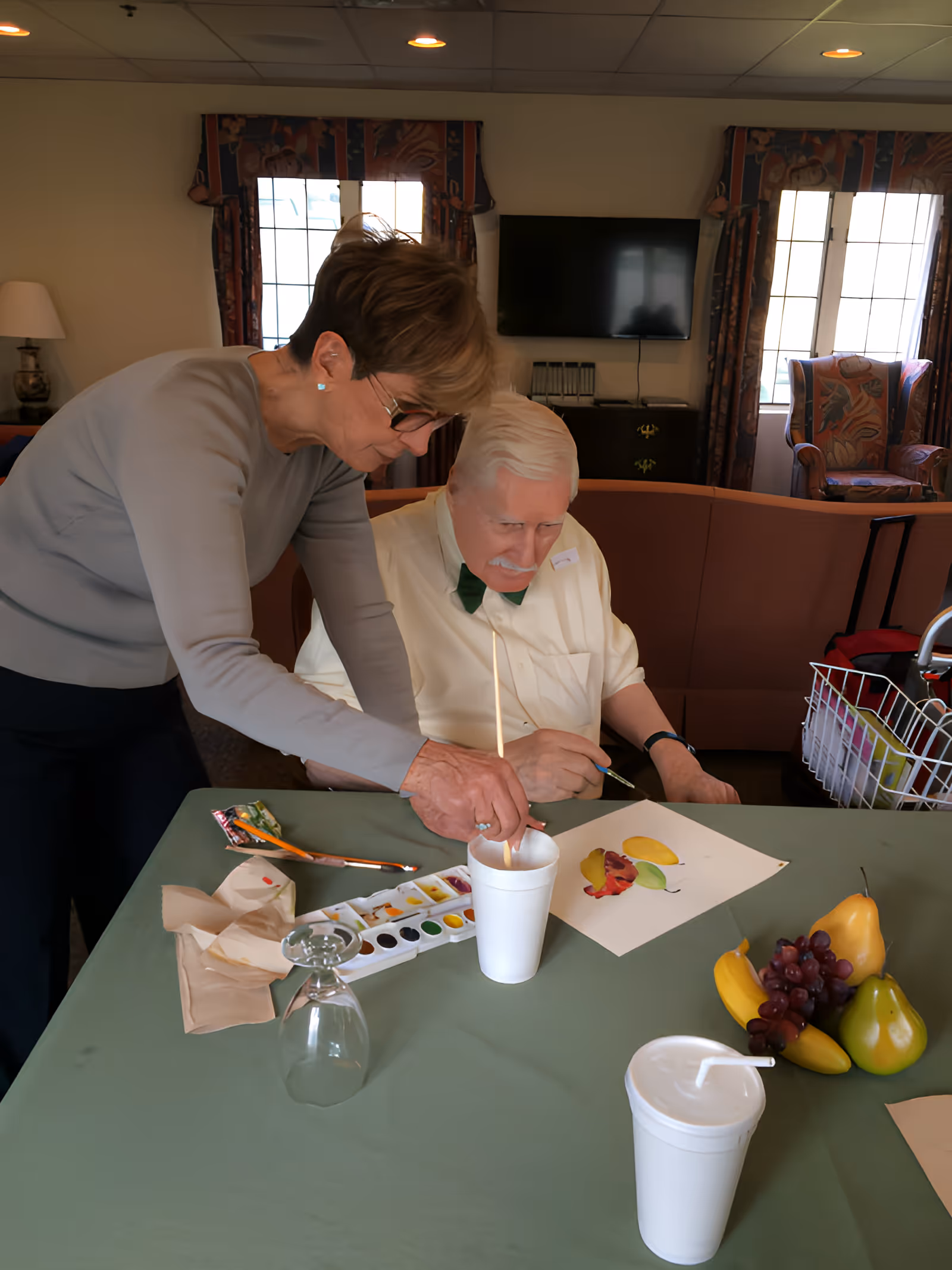 An elderly man seated at a table is painting a picture of fruit with watercolors while a woman stands beside him, assisting by holding a cup of water. The table is covered with a green tablecloth and has painting supplies, a glass, a white cup with a straw, and a small arrangement of artificial fruit. The room has a cozy interior with a sofa, armchairs, windows with patterned curtains, and a wall-mounted TV in the background.