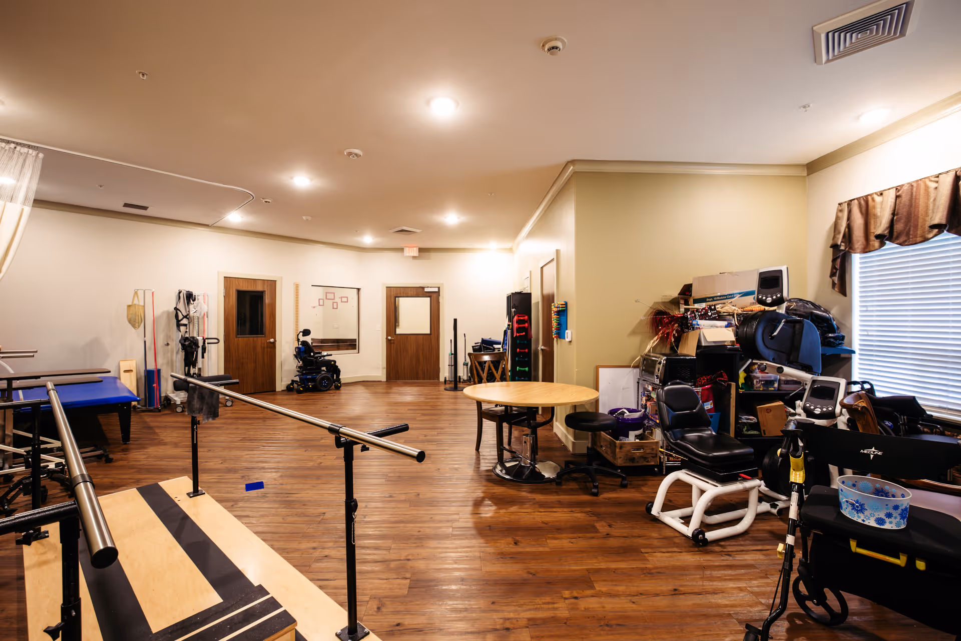 Interior view of a rehabilitation room with wooden floors and beige walls. The room contains various physical therapy equipment including parallel bars, exercise tables, wheelchairs, and other medical devices. There is a round table with stools near the center and a window with blinds and a valance on the right side.