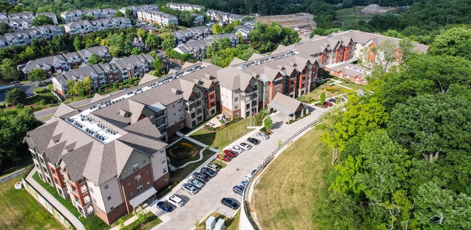 Aerial view of a large senior living facility named Harmony at Brentwood, surrounded by trees and greenery. The building is multi-story with a mix of beige and red exterior walls and a gray roof. There is a parking lot with several cars parked in front of the building, and residential houses are visible in the background.