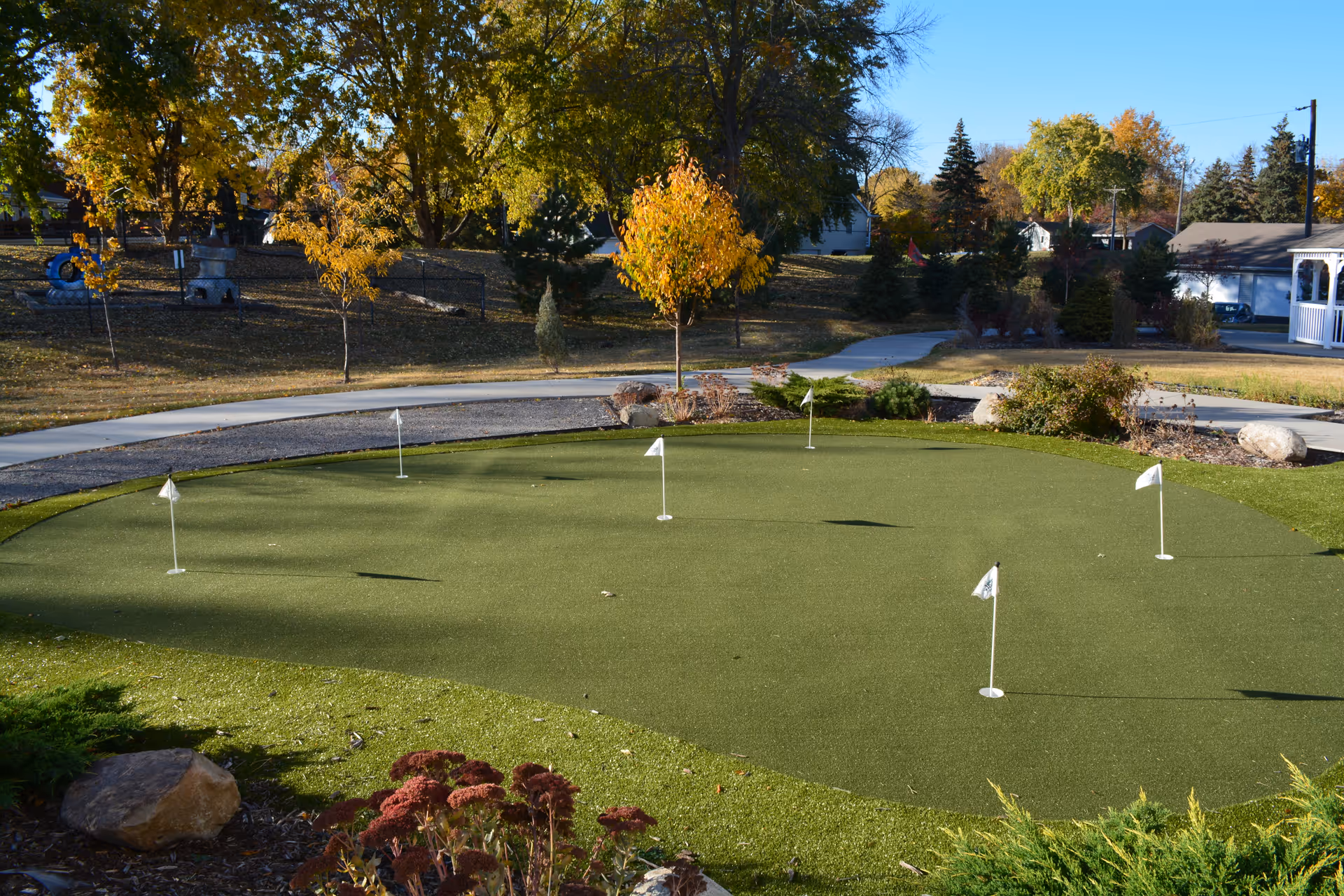 Outdoor putting green with several small white flags on a sunny day, surrounded by trees with autumn foliage, shrubs, rocks, and a paved walking path in the background.