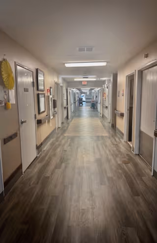 A long, well-lit hallway in a senior living facility with wood-patterned flooring, beige walls, and multiple doors on both sides. Handrails run along the walls, and there are framed pictures and decorations on the left side. The hallway leads to a brighter area in the distance.