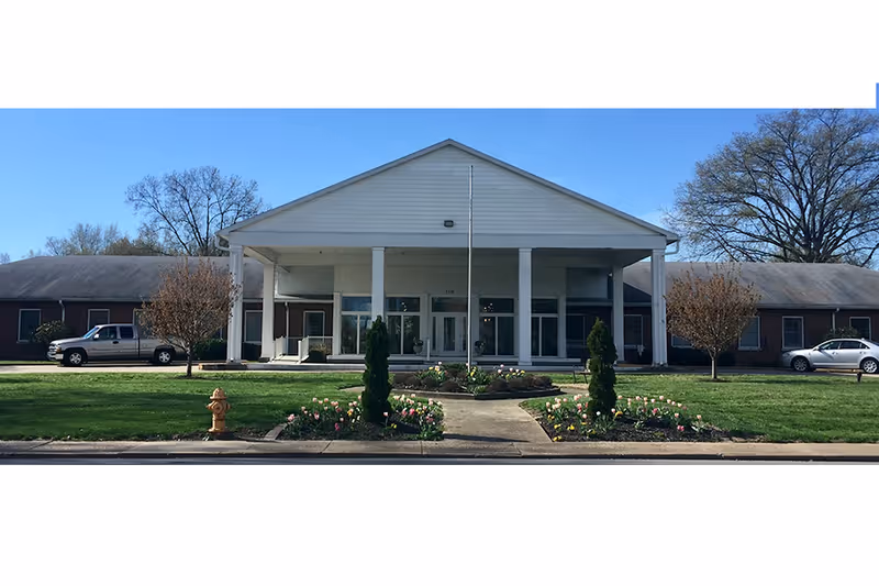 Front exterior view of a single-story building with a large covered entrance supported by white columns, surrounded by a well-maintained lawn, flower beds, and trees. Two vehicles are parked on either side of the building under a clear blue sky.