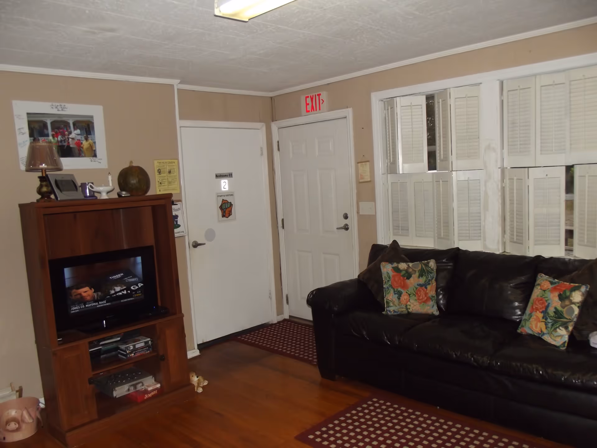 Interior view of a living room with a black leather couch adorned with floral pillows, a wooden entertainment center with a television, and a door marked as Restroom #2. The room has beige walls, white window shutters, and a wooden floor with patterned rugs. An exit sign is visible above a white door.
