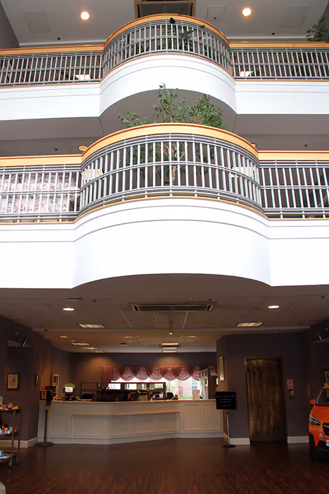 Interior view of a senior living facility lobby with a white reception desk, purple walls, and a wooden floor. Above the lobby area, there are two curved balconies with white railings and some greenery visible. The ceiling has recessed lighting and air vents.