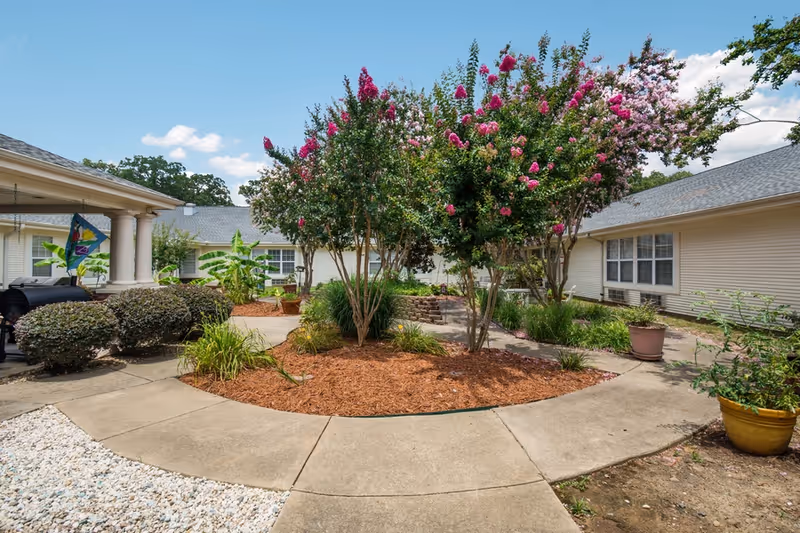 Outdoor courtyard area with a circular concrete walkway surrounding a mulched garden bed with blooming pink and white flowering trees. The courtyard is bordered by single-story beige buildings with windows, and there are potted plants and shrubs along the walkway. The sky is clear with a few clouds.