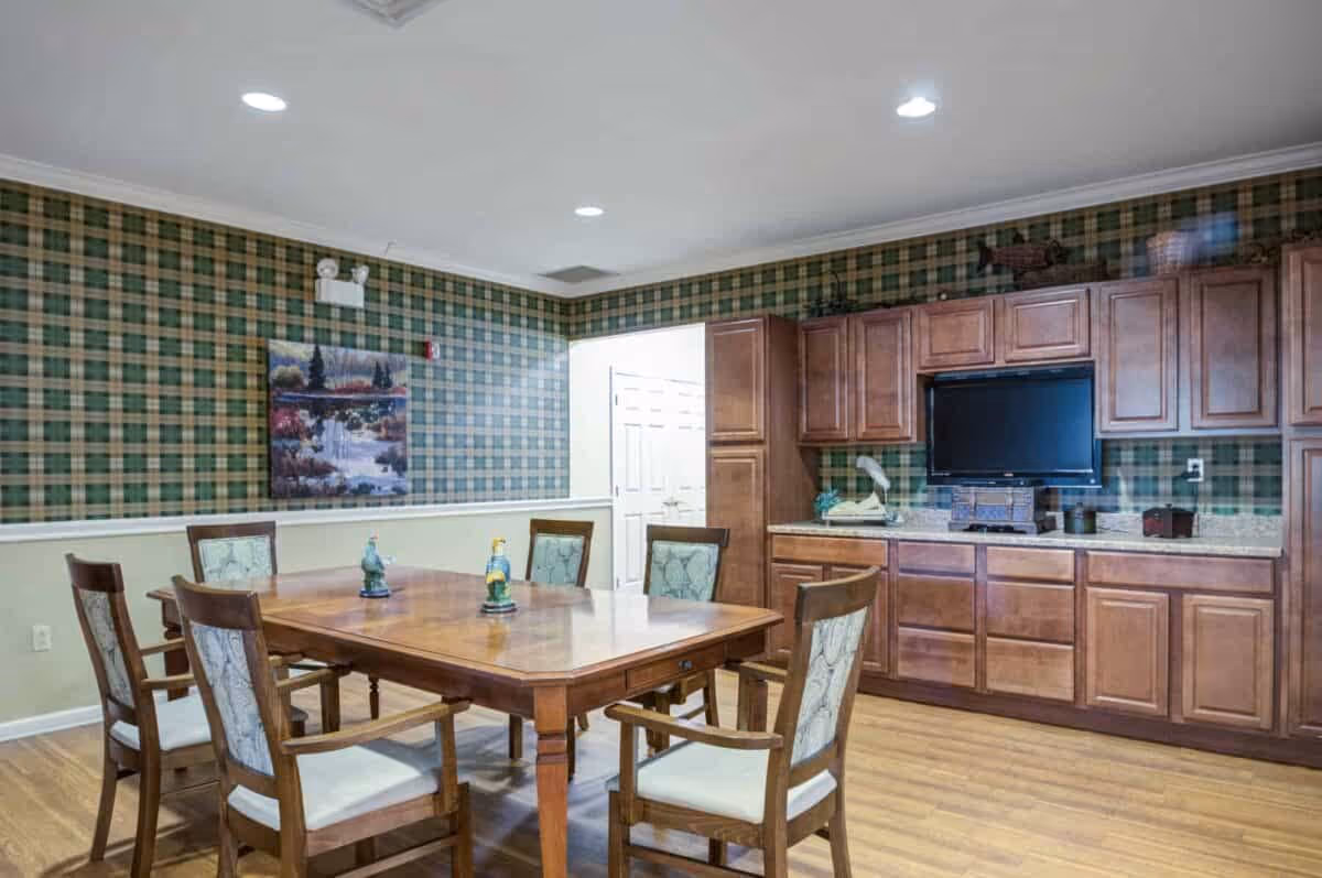 A room with a wooden table surrounded by six wooden chairs with cushioned seats and backs. The walls have green and beige plaid wallpaper on the upper half and a beige paint on the lower half. There is a painting of a waterfall on one wall. On the right side, there are wooden cabinets with a countertop and a mounted flat-screen TV above it. The floor is wooden, and the ceiling has recessed lighting.