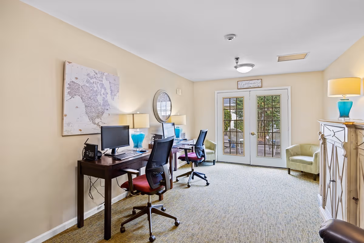 A bright and cozy office area in a senior living facility with two desks, each equipped with a computer and a turquoise lamp. Two office chairs with red seats are positioned at the desks. The room has beige walls, a carpeted floor, a large wall map, a round mirror, and a pair of French doors leading outside. There are two green armchairs near the doors and a wooden cabinet with another turquoise lamp on top.