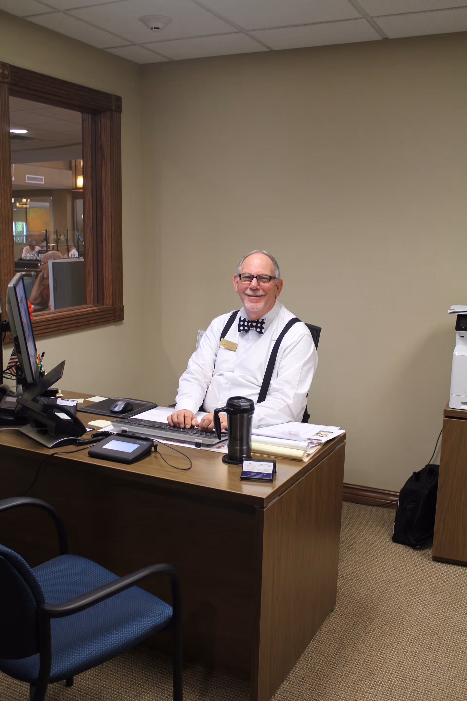 An elderly man wearing glasses, a white shirt, black suspenders, and a polka dot bow tie is sitting at a wooden desk in an office. He is smiling and typing on a keyboard. The desk has a computer monitor, a phone, a travel mug, papers, and office supplies. There is a blue chair in front of the desk and a window with a wooden frame showing another room in the background.