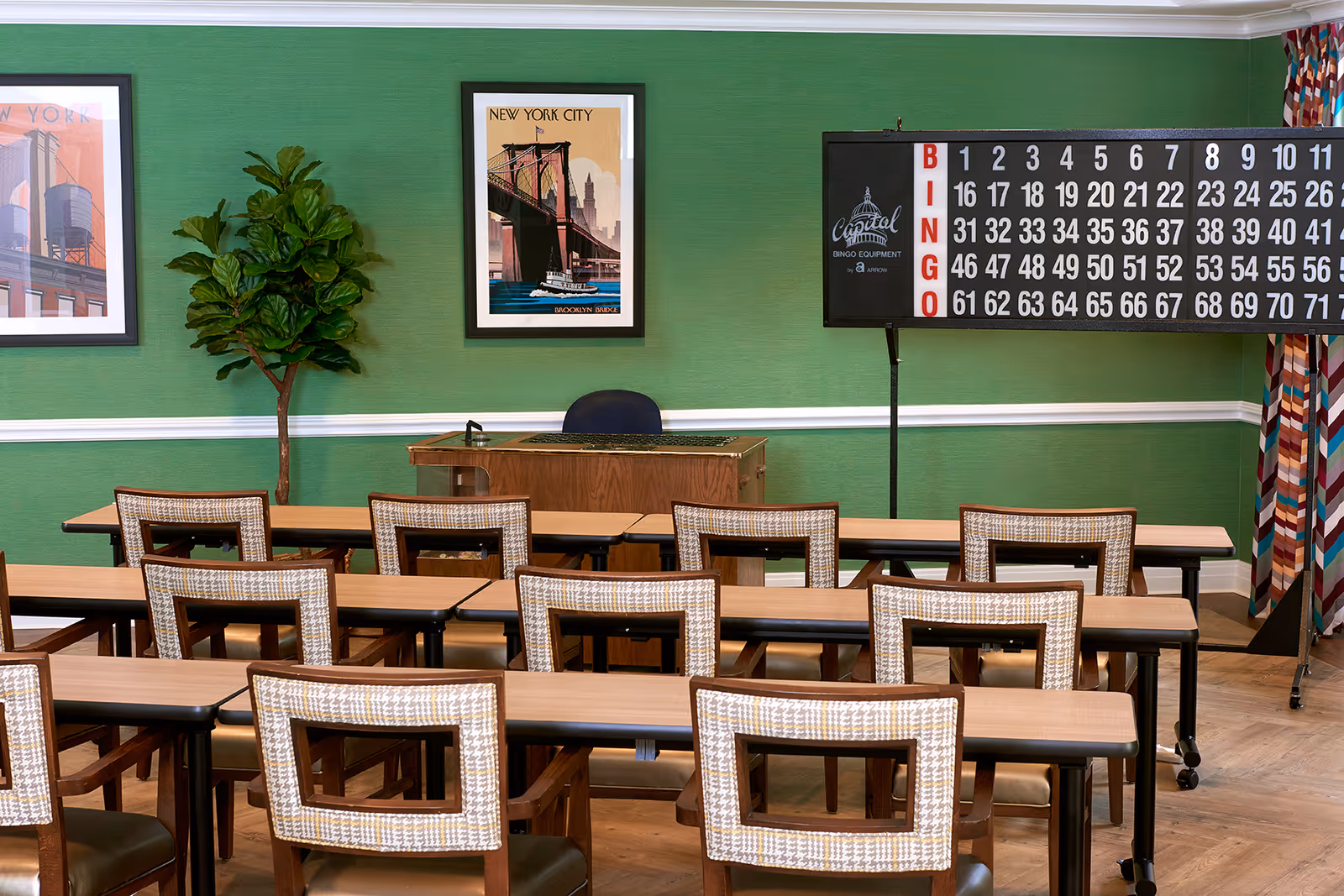 A bingo room with rows of wooden tables and chairs facing a wooden desk and a large bingo board on a stand. The walls are green with white trim, decorated with framed pictures of New York City and a potted plant in the corner.