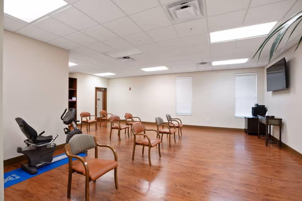 A spacious room with wooden flooring and beige walls, featuring several chairs arranged in rows, an exercise bike on a blue mat, a wall-mounted TV, and a small table with electronic equipment near two windows with blinds.