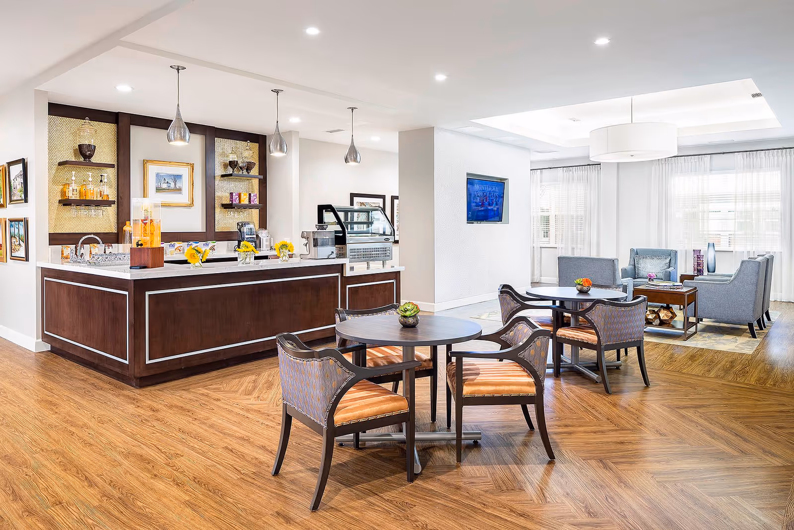 A bright and modern common area in a senior living facility featuring a wooden floor, a coffee and snack bar with pendant lights, round tables with chairs, and a seating area with sofas and a TV mounted on the wall.