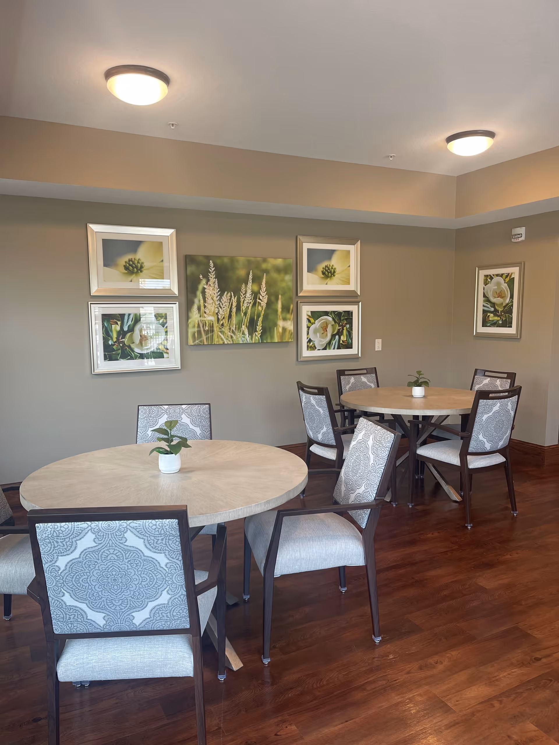 A dining area with two round wooden tables, each surrounded by four cushioned chairs with patterned backs. The room has wooden flooring and beige walls adorned with framed floral and nature artwork. Two ceiling lights illuminate the space.