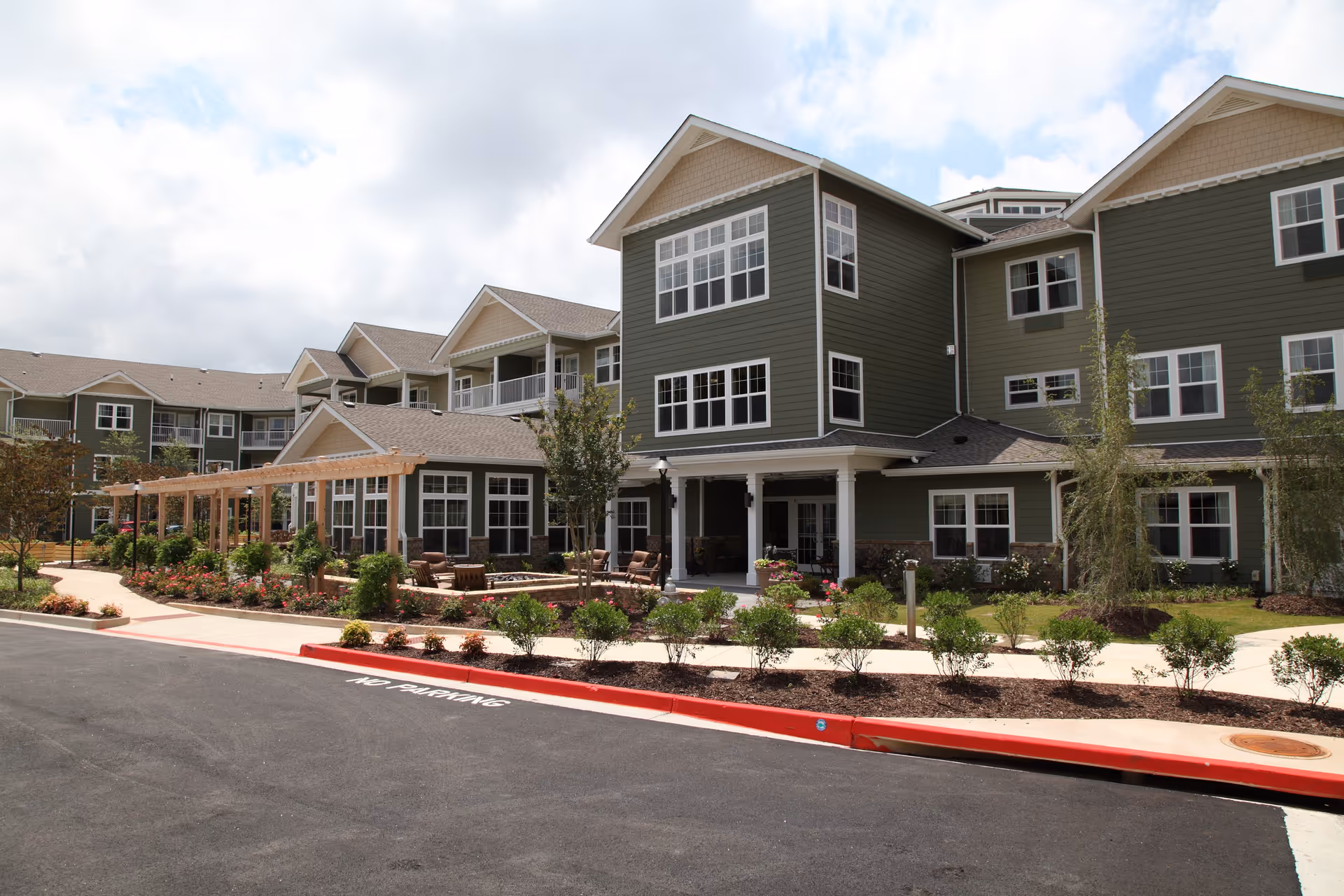 Exterior view of a multi-story senior living facility with green siding and white trim. The building features large windows, a covered entrance, and a landscaped garden with shrubs and flowers. There is a paved driveway with a red curb marked 'NO PARKING'.