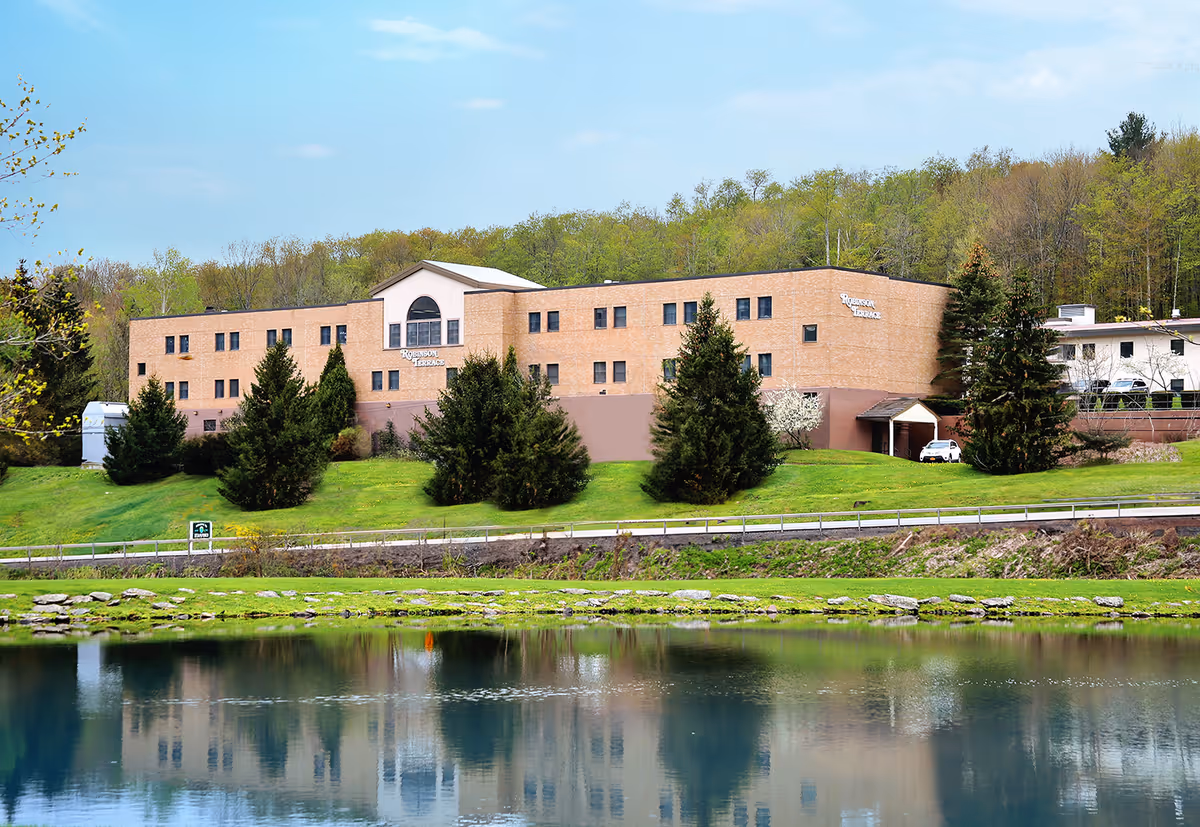 A three-story brick rehabilitation and nursing center on a grassy hill with trees, reflected in a pond in the foreground.