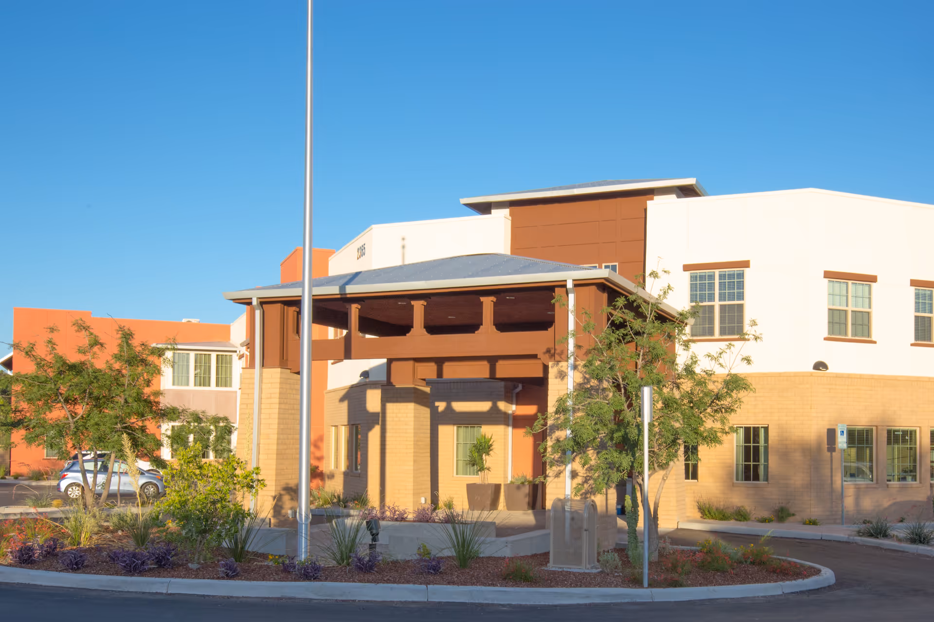 Exterior view of The Ranch Estates of Tucson building entrance with a covered porch, surrounded by landscaping and trees under a clear blue sky.