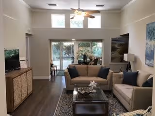 A bright living room with two beige sofas, a glass coffee table, a patterned rug, and a wooden TV stand with a flat-screen TV. Large windows and glass doors at the back let in natural light and offer a view of an outdoor patio area. The room has a ceiling fan with lights and neutral-colored walls with artwork.