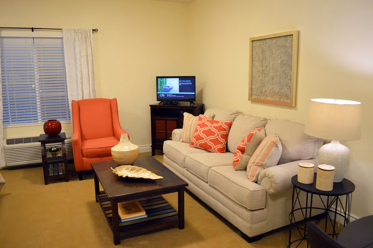 A cozy living room with a beige sofa adorned with red and striped pillows, a coral armchair, a dark wooden coffee table with decorative items, a side table with a lamp and two small containers, a small TV on a stand, and a window with white blinds and curtains.