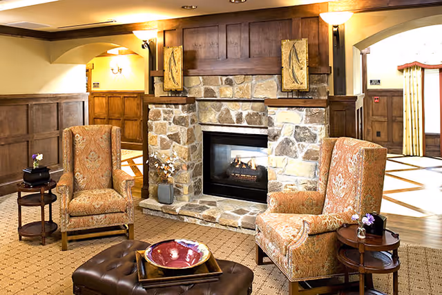 A cozy living room area featuring a stone fireplace with a wooden mantle, two patterned armchairs, a brown leather ottoman with a decorative bowl on top, and small side tables with flowers. The room has warm lighting and wood-paneled walls.