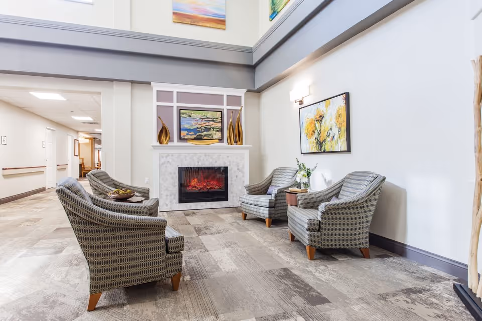 A bright and spacious sitting area in a senior living facility with four patterned armchairs arranged around an electric fireplace. The fireplace is decorated with gold vases and a painting above it. There is a colorful floral painting on the wall to the right and a small side table with a flower arrangement. The floor is carpeted with a modern pattern, and the walls are painted white with gray trim. A hallway is visible to the left.