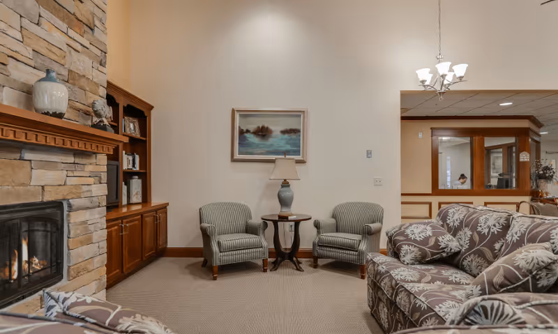 A cozy living room area with a stone fireplace on the left, a wooden built-in bookshelf, two striped armchairs with a small round table and lamp between them, and a floral patterned sofa in the foreground. A framed painting hangs on the wall above the armchairs, and a chandelier is visible in the adjacent room.