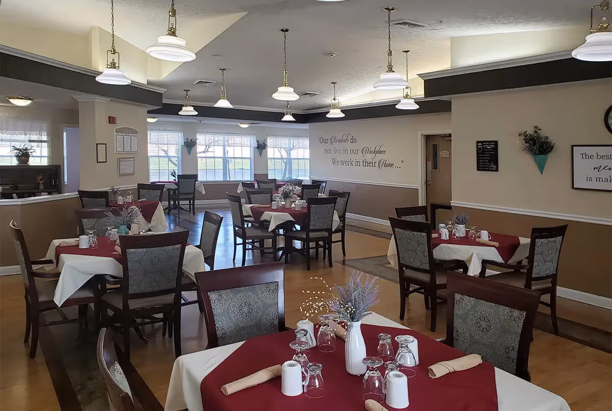 A dining room with multiple tables covered with white and burgundy tablecloths, each set with glasses, napkins, and a small vase with flowers. The room has large windows letting in natural light, pendant lights hanging from the ceiling, and inspirational wall quotes.