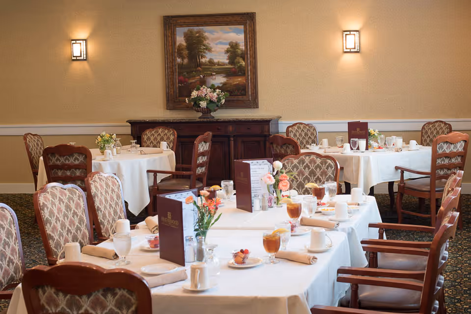 Dining room with multiple white-clothed tables set with glasses, plates, menus, and floral centerpieces in an elegant dining area.