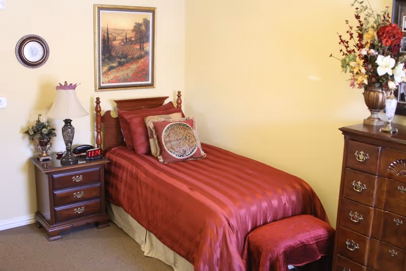 A cozy bedroom with a single bed covered in a red striped bedspread and decorative pillows. Next to the bed is a wooden nightstand with a lamp, a clock, and a small plant. Above the bed hangs a framed landscape painting. On the right side, there is a wooden dresser with a floral arrangement on top. The walls are painted light yellow.