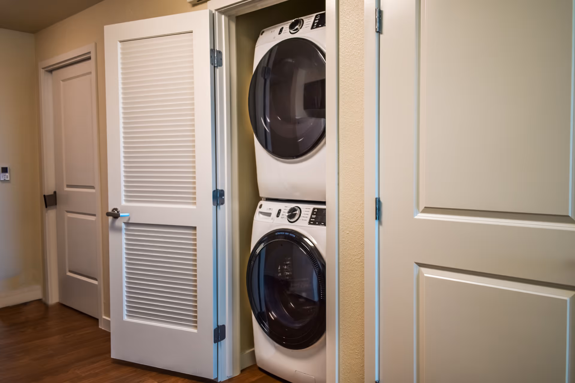 A laundry closet with a stacked washer and dryer inside. The closet has white louvered doors, one of which is open to reveal the appliances. The surrounding walls are beige, and there are two closed doors nearby with silver handles. The floor is wooden.