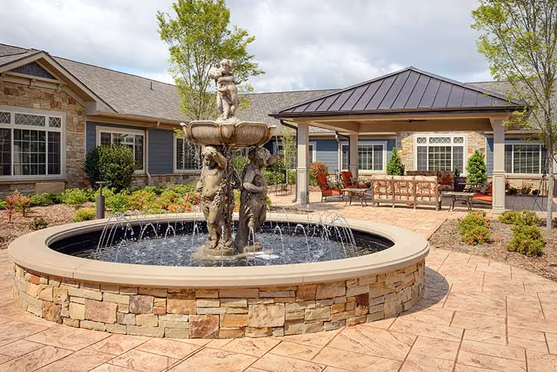 Outdoor courtyard area with a stone fountain featuring three cherub statues and water jets. Surrounding the fountain is a stone-paved patio with a covered seating area furnished with chairs and a sofa. The background shows a building with stone and siding exterior, windows, and some landscaping with trees and shrubs under a partly cloudy sky.