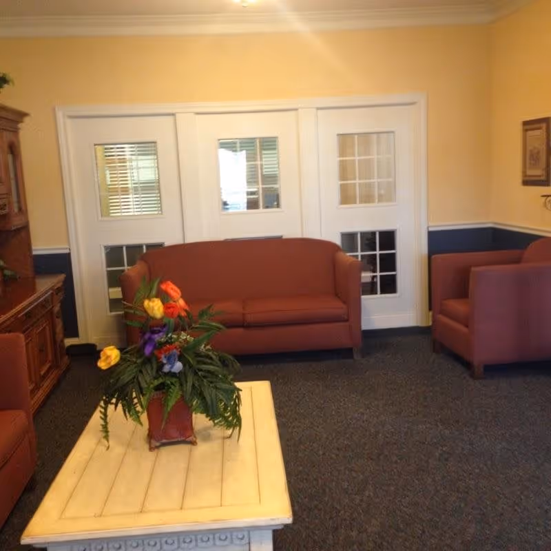 A cozy living room area with a brown sofa and matching armchair, a white wooden coffee table with a colorful flower arrangement, and a wooden cabinet against a yellow and blue wall with white framed windows.