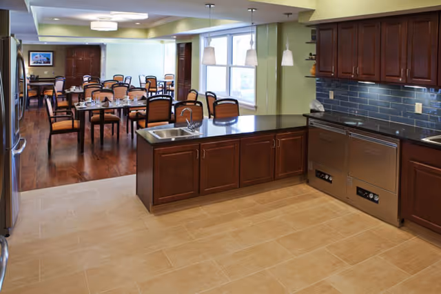 Open communal kitchen with a center island and sink overlooking a dining room set with multiple tables and chairs.